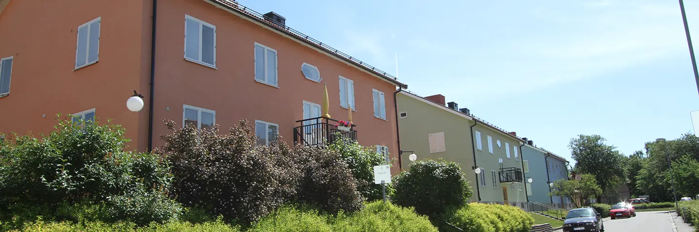 Tre tvåvåningshus med putsade fasader i orange, grön och blå. Grönska och villagata i förgrund. 