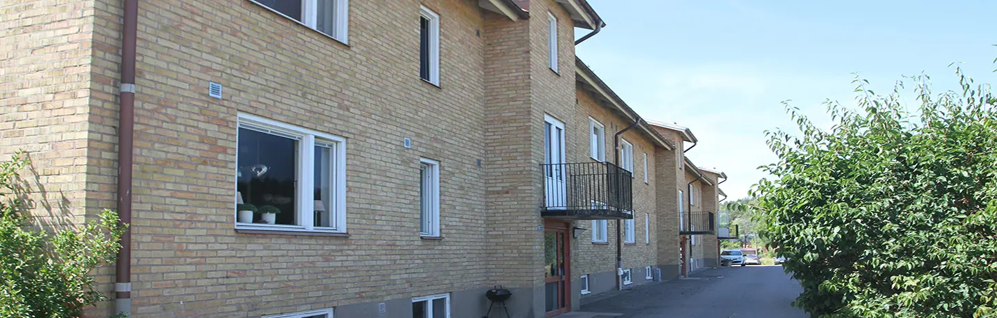 Fasadbild över kvarteret Fors i Sjuntorp. Tegelfasad med vitt runt fönster, svarta balkonger, buskar i förgrund.