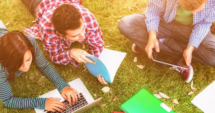 Tre personer sitter i gräset och studerar framför skärmar och block