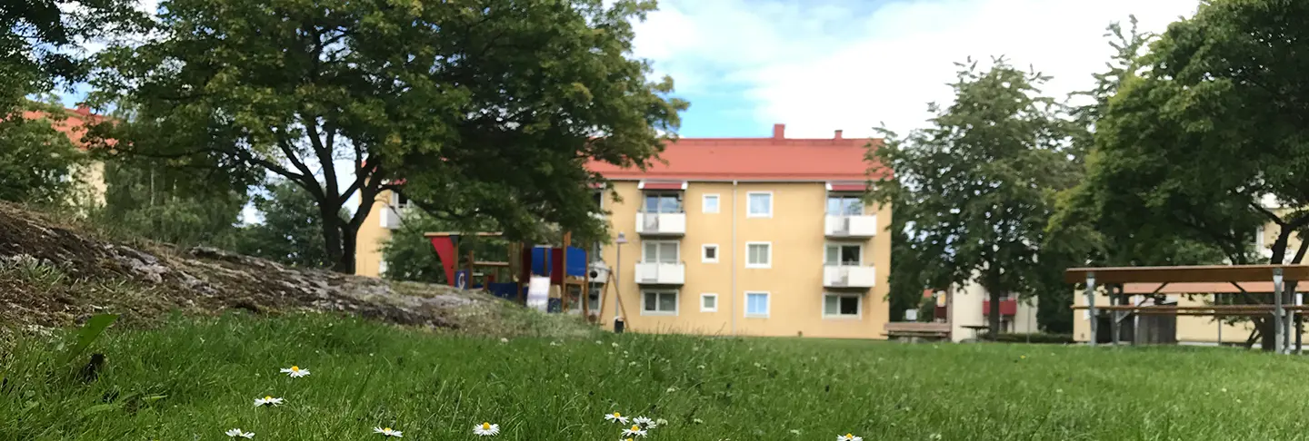 Fasadbild över kvarteret Beckasinen i Trollhättan. Gul fasad med vita balkonger, i förgrund grönska och lekplats